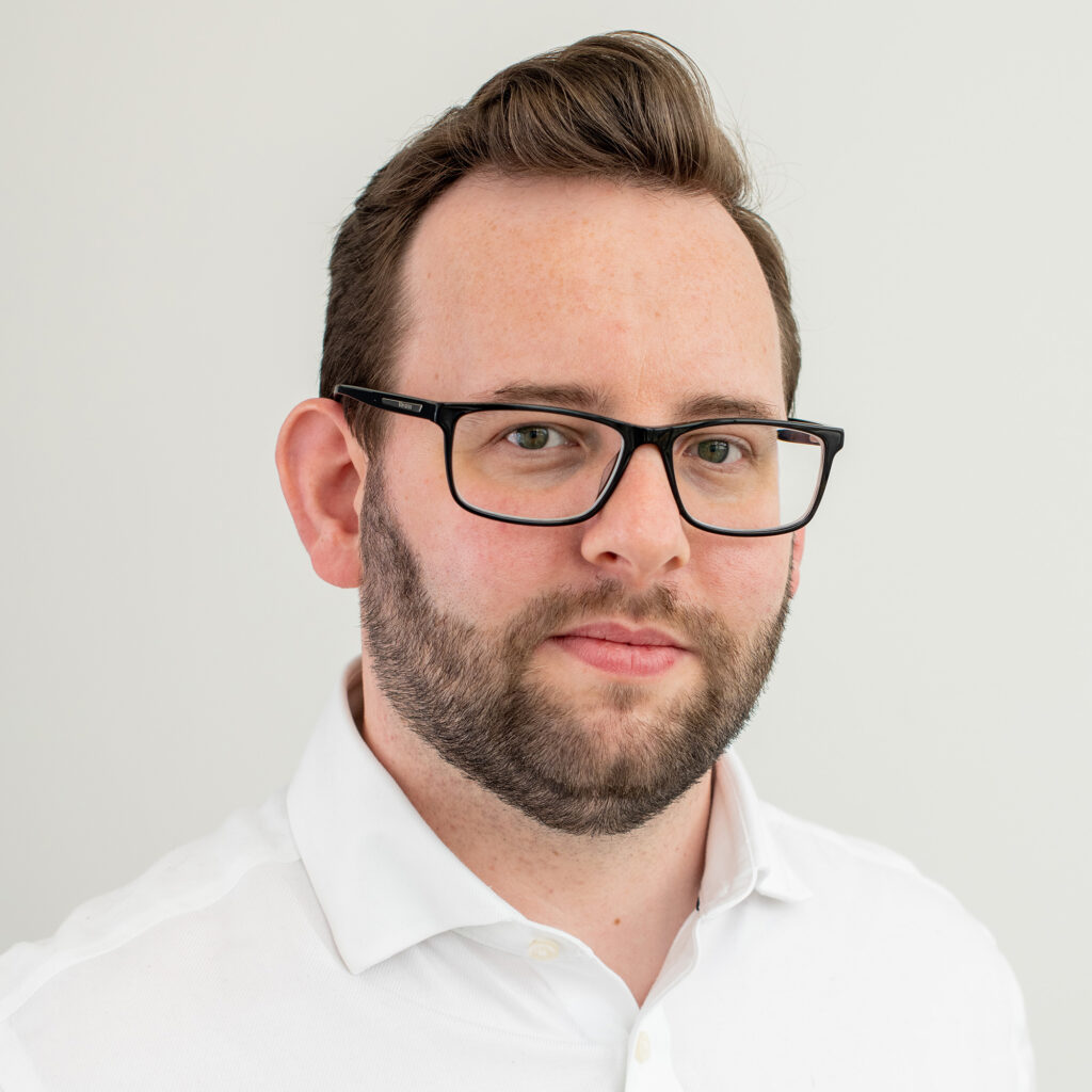 A headshot of David Molloy, a 30-something man with a short cropped beard and black glasses, wearing a white work shirt.